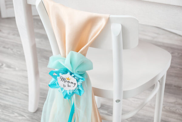 White chair decorated with peach and aqua fabric sash and a "Mommy to Be" rosette for a baby shower