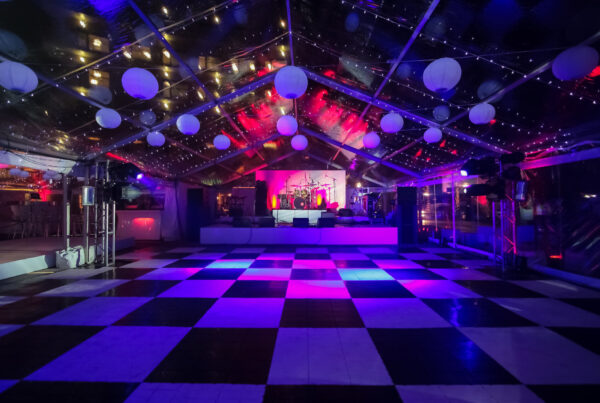 Black and white checkered dance floor in a clear-roof event tent with paper lanterns and a lit stage