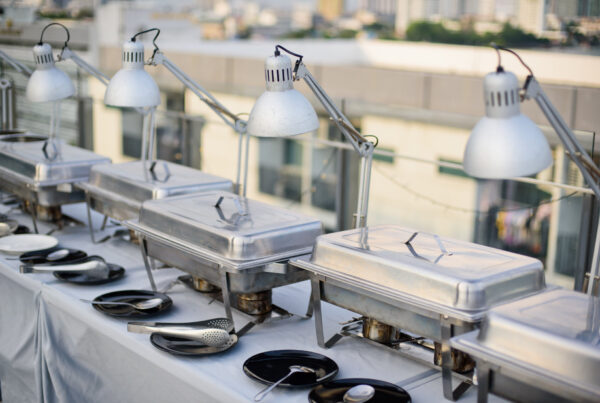 Row of stainless steel chafing dishes with heat lamps set up on a rooftop party buffet table