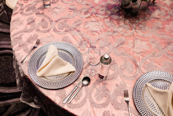 Round event table dressed in a pink damask-patterned tablecloth with silver charger plates, folded napkins and glassware