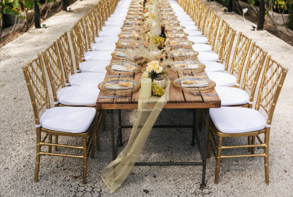 Long rustic wooden banquet table lined with gold chiavari chairs set for an outdoor event