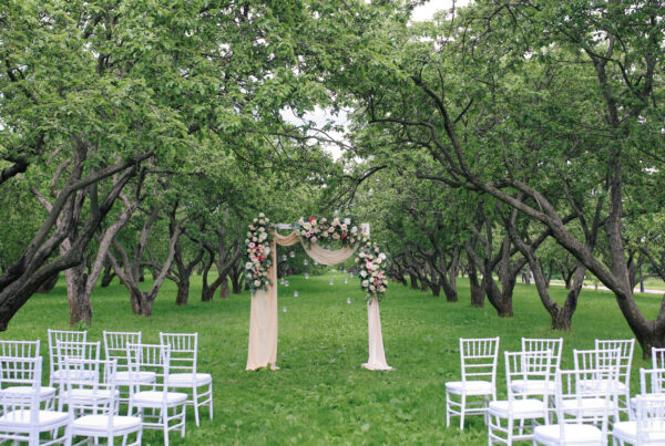Outdoor wedding ceremony with a floral draped arch and white chairs on a lawn beneath trees