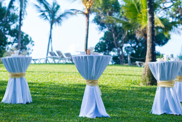 Tall cocktail tables draped in white linens with gold sashes on a lawn at an outdoor event