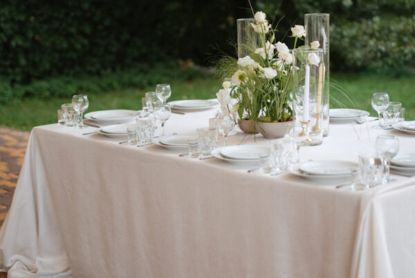 Ivory rectangle tablecloth draped to the floor on an outdoor wedding reception table with white place settings and floral centerpiece