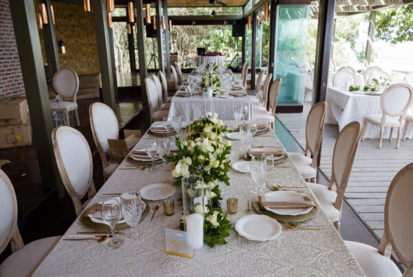 Long rectangular banquet table set with white plates, glassware and floral centerpieces, lined with upholstered chairs at a wedding reception